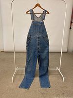 Full front view of vintage Lee denim overalls hung on hanger and rack, showing the overall shape and condition.