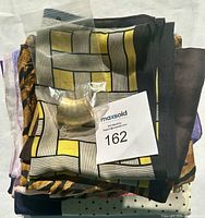 Folded group of silk scarves in different colors and patterns with a small gold-toned crescent brooch in a plastic bag on top