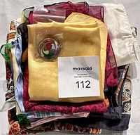 A stack of various silk scarves in different colors and patterns, some sheer and solid, with a yellow scarf prominently on top and a decorative pin/brooch placed on it.