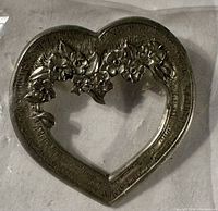 Metallic heart-shaped brooch pin or brooch with floral embossed design on top and side edges.