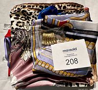 Folded variety of 100% silk scarves in multiple colors and patterns. Includes a small gold-tone pin or brooch nestled in the scarves. MaxSold lot tag included in photo.