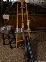 Overall view of art supplies including wooden easel, artist mannequin, books, drawing tubes, storage containers on a wooden stool with removable cushion