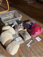 Photo showing assorted colored wool batts and packets including natural white, black, grey, purple, red, and pink shades spread out on wooden floor beside a clear plastic storage bin and needle holder.