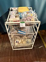 White wire metal storage bin with three pull-out wire baskets filled with assorted materials including yarn, crochet needles, and crafting accessories.