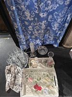 Full lot view showing scarves, trays, glassware, and decorative items on black table with floral curtain background.