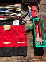 Photo shows multiple knitting needles in various sizes and materials arranged with a red and black roll-up needle pouch and plastic containers containing smaller tools.