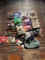 Overview photo showing multiple plastic bags of yarn in assorted colors and textures, stacked and arranged with books and needle accessories.