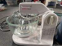 Sunbeam Mixmaster hand mixer on a white base with two nested glass mixing bowls placed inside the larger bowl, power cord wrapped around mixer.
