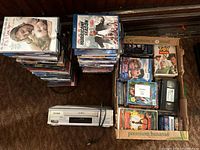 Photo showing stacks of DVDs, a Panasonic cassette player with power cord, and two boxes containing assorted VHS and DVDs.