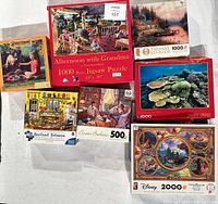 Photo displaying 7 jigsaw puzzles with various themes and piece counts. Boxes are placed on a white surface showing titles and images.