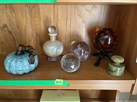Photo of six art glass items on a wood shelf including paperweights, glass globe on stand, and decanter bottle.