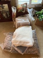 Full view of lot showing decorative floral pillow, pack of linens, white plush blanket, and additional textured bedding pieces on floor.