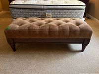 Front view of brown upholstered tufted bench seat with wooden legs beside a mattress.