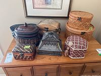 Photo showing eight decorative woven and metal keepsake boxes and baskets arranged on wooden table under framed picture.