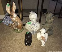 Five assorted figurative sculptures on carpeted floor: ceramic rabbit, Cupid, white bust, black abstract figure, and gray winged fairy.