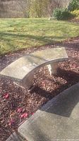 Photo of curved concrete garden bench placed outdoors on mulch bedding with grass and sidewalk in background.