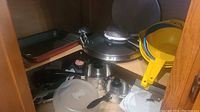 Overview of various pots, pans, lids, and yellow plastic colanders inside a wooden cabinet shelf.