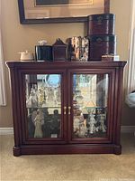 Front view of wooden display cabinet with glass doors and decorative items on top. Shows two doors with brass handles and glass panels.
