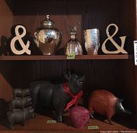 Shelf showing two wooden ampersand signs, large silver mercury glass jar with lid, art glass bottle with bird topper on top shelf. Lower shelf has cast iron pig figurines (large with red ribbon and three small stacked), carved wood pig figurine.