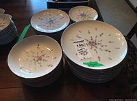Multiple stacks of white china plates with small pink and purple rosebud floral designs and green leaves in varied sizes.
