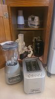 Wide view showing Oster blender, Breville toaster, and various food choppers and mixer attachments inside wooden cabinet.