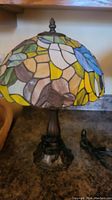 Full view of the stained glass style table lamp with colorful acrylic shade and dark metal base on a countertop.