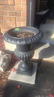 Front-right angled view of black cast iron planter urn filled with soil, showing molded decorative details and pedestal base.