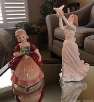 Front view of both Royal Doulton figurines on a reflective surface shown with background couch and room light