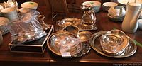 Wide view of multiple silverplated serving ware pieces on wood table including dishes, bowls, utensils, and trays.