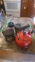 Red Le Creuset tea kettle, two coffee carafes with black lids, and metal racks grouped on kitchen counter.
