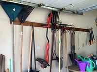 Wide view showing blue and black leaf rakes, red electric edger, pitch forks, shovels, and other tools hanging on garage wall