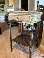 Antique wooden side table with pale green distressed finish, one drawer with brass round knob, and cane shelf below.