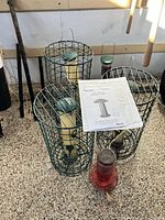 Five bird feeders including three green metal cage caged tube feeders, one red hummingbird feeder, and one new feeder, arranged on gravel with light.