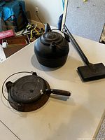 Three cast iron pieces on table showing kettle, waffle iron, and bed warmer with varying shapes and sizes