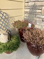 Front view of two large brown ceramic planter pots with plants, one with dried foliage and the other with greenish succulents, a smaller resin pot with green foliage, two metal trellises behind large pots, and one small white owl statue inside a pot