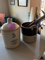 Photo showing two large crocks, one beige with a handle and the other two-tone black and cream with wooden sticks inside.