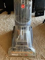 Front view of Hoover MaxExtract77 carpet cleaner showing transparent water tank and Hoover logo on a carpeted floor.