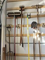 Full view of rakes, push brooms, shovel, and pothole diggers hanging on wall