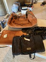 Overview showing large tan leather duffel bag open on top, brown leather portfolio beside it, black nylon zippered bag in front, and black drawstring nylon cosmetic bag to right.