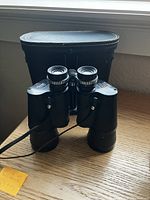 Black vintage Manon 10x50 binoculars positioned upright with black carrying case in background on wooden surface.