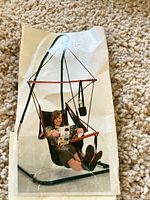Photo of the original product image showing a woman seated in the fully assembled hammock chair with metal stand and wooden supports, reading a magazine.