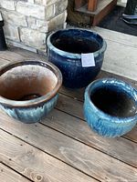 Three planter pots shown together on wooden deck against stone wall - two dark blue ceramic planters, one smaller blue-green resin pot visible.