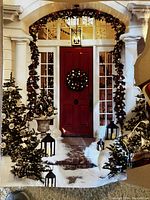 Decorative pre-lit holiday garland wrapped around exterior doorframe with lit small evergreen trees and lanterns around door, showing use scenario.