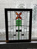 Frontal image of stained glass panel showing orange flower and green stem with clear textured glass background and wooden frame.