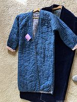 Two Uzbekistani men's robes shown on hangers and carpeted floor: one blue quilted with patterned lining, one dark and velvet with floral cuff detail.