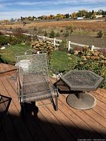 One rocker chair with adjacent hexagonal metal mesh table on wooden deck with outdoor plants and field background