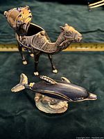 Metallic camel figurine with intricate engravings, blue stone inlays, and open compartment on its back, positioned next to dolphin figurine with rhinestones.