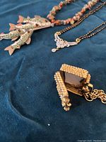 Close-up photo showing the open gold-tone suitcase locket pendant, the rhinestone-decorated chain section, and the three fish-shaped charms with multicolored stone chip beads.