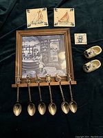 Full view of the lot on black background showing the Delft hanging tile, five spoons, two small coasters, and pair of wooden shoes.