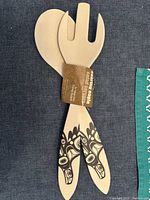 Pair of bamboo salad servers shown laid side by side on fabric background, with attached product label identifying design and artist.